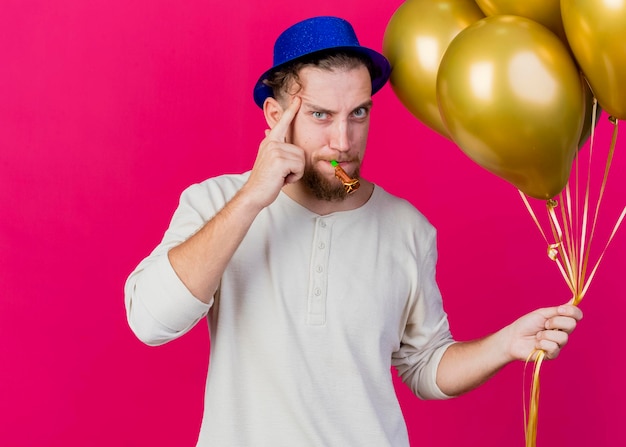 Confident young handsome slavic party guy wearing party hat holding balloons and party blower looking at front doing think gesture isolated on pink wall