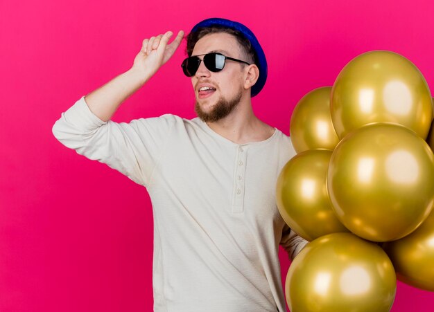 Confident young handsome slavic party guy wearing party hat and sunglasses holding balloons looking at side doing loser gesture showing tongue isolated on pink wall