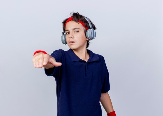 Confident young handsome sporty boy wearing headband and wristbands and headphones with dental braces looking and pointing at camera isolated on white background with copy space