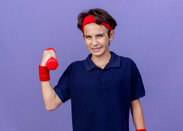 Confident young handsome sporty boy wearing headband and wristbands with dental braces  holding dumbbell isolated on purple wall with copy space