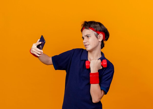 Confident young handsome sporty boy wearing headband and wristbands with dental braces holding dumbbell taking selfie isolated on orange wall