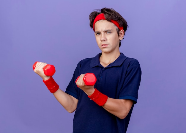 Confident young handsome sporty boy wearing headband and wristbands with dental braces  holding dumbbells isolated on purple wall with copy space