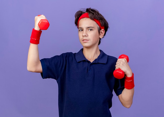 Confident young handsome sporty boy wearing headband and wristbands with dental braces holding dumbbells looking at front isolated on purple wall
