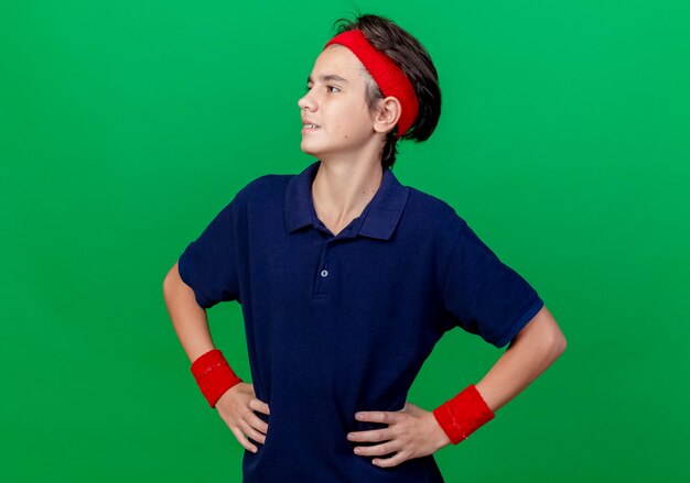 Confident young handsome sporty boy wearing headband and wristbands with dental braces keeping hands on waist looking at side isolated on green wall