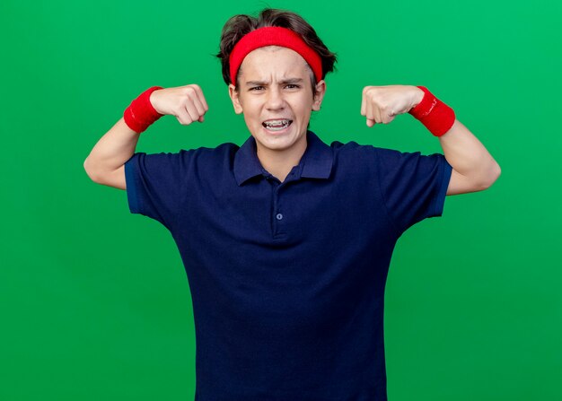 Confident young handsome sporty boy wearing headband and wristbands with dental braces looking at front doing strong gesture isolated on green wall