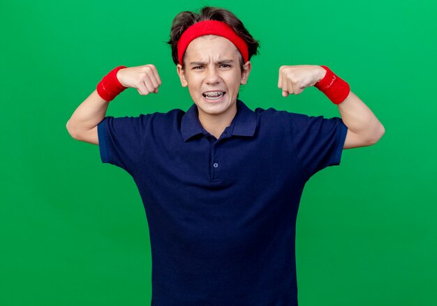 Confident young handsome sporty boy wearing headband and wristbands with dental braces looking at front doing strong gesture isolated on green wall