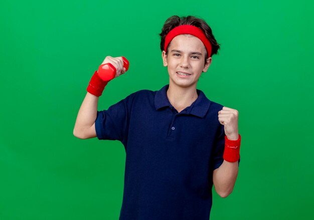 Confident young handsome sporty boy wearing headband and wristbands with dental braces looking at front raising up dumbbell doing yes gesture isolated on green wall with copy space