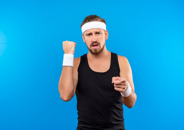 Confident young handsome sporty man wearing headband and wristbands clenching fist and pointing  isolated on blue wall with copy space