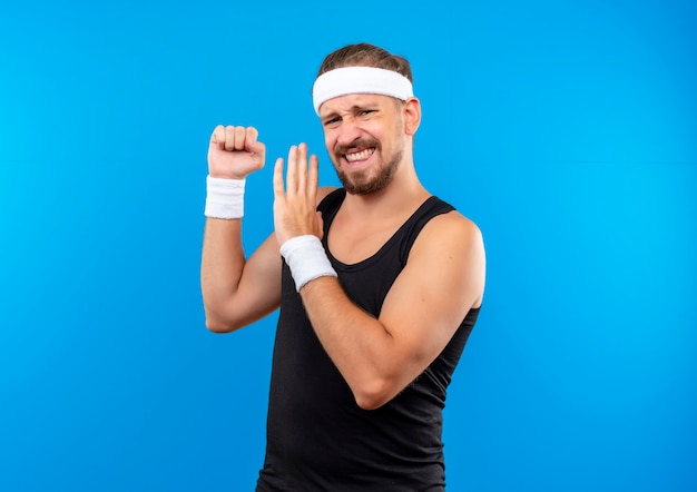 Уверенный молодой красивый спортивный мужчина с головной повязкой и браслетами, сжимая кулак и указывая рукой на него, изолированную на синей стене с копией пространства