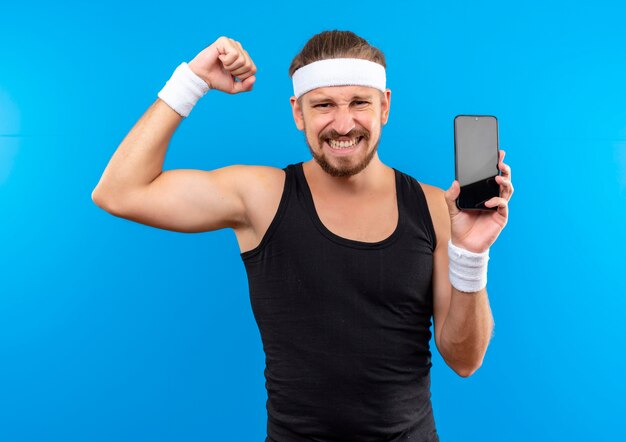 Confident young handsome sporty man wearing headband and wristbands gesturing strong and holding mobile phone isolated on blue wall