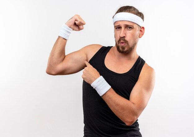 Confident young handsome sporty man wearing headband and wristbands gesturing strong and pointing at his muscles isolated on white wall