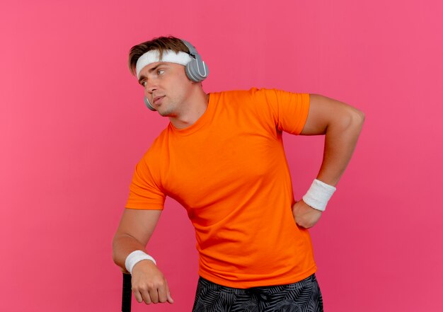 Confident young handsome sporty man wearing headband and wristbands and headphones putting hand on waist and another one on baseball bat looking at side isolated on pink  with copy space