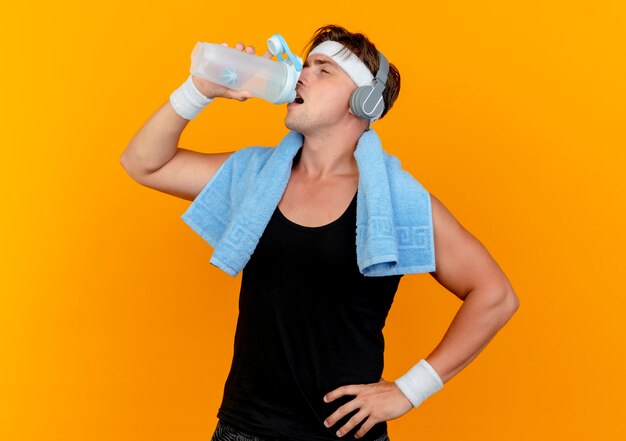 Confident young handsome sporty man wearing headband and wristbands and headphones with towel around neck drinking water from bottle with hand on waist isolated on orange 