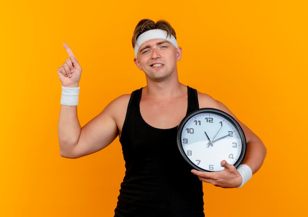 Confident young handsome sporty man wearing headband and wristbands holding clock and pointing up isolated on orange 