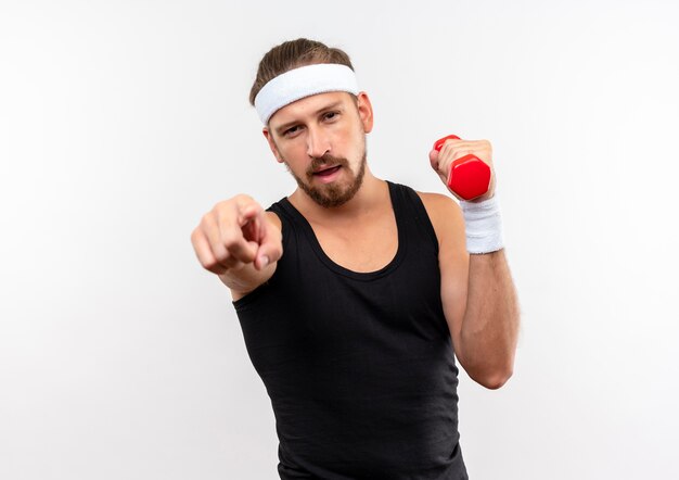 Confident young handsome sporty man wearing headband and wristbands holding dumbbell and pointing  isolated on white wall