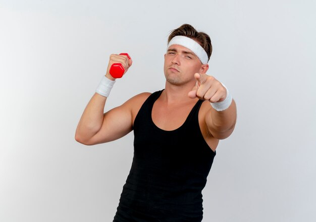 Confident young handsome sporty man wearing headband and wristbands holding dumbbell and pointing isolated on white  with copy space