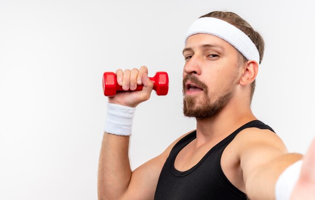 Confident young handsome sporty man wearing headband and wristbands holding dumbbell and stretching out hand  isolated on white wall with copy space