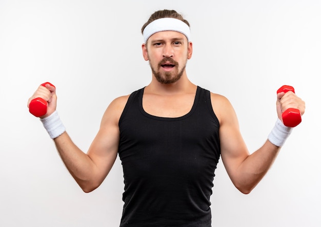 Confident young handsome sporty man wearing headband and wristbands holding dumbbells and looking  isolated on white wall