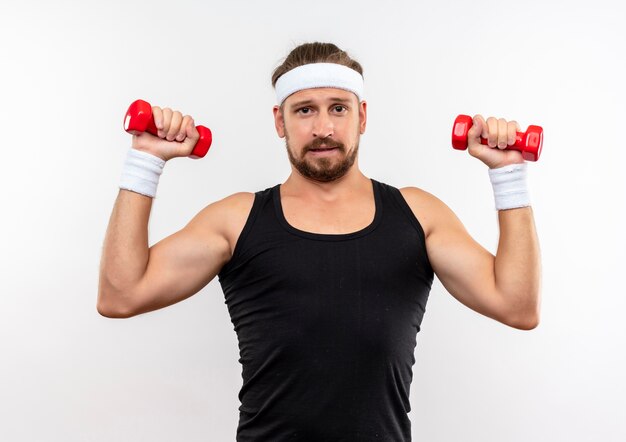 Confident young handsome sporty man wearing headband and wristbands holding dumbbells looking  isolated on white wall
