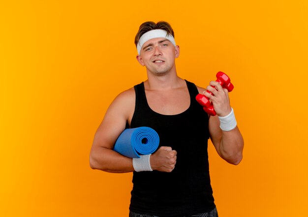 Confident young handsome sporty man wearing headband and wristbands holding dumbbells with towel isolated on orange  with copy space