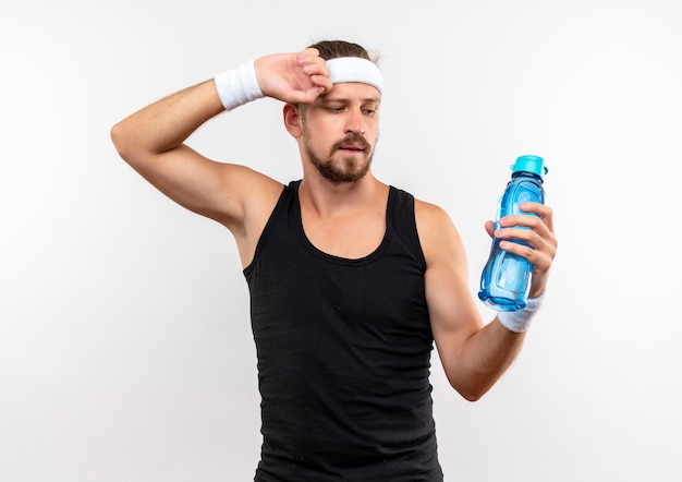 Confident young handsome sporty man wearing headband and wristbands holding and looking at water bottle with hand on head isolated on white wall