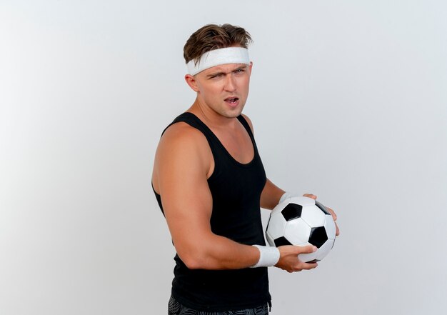 Confident young handsome sporty man wearing headband and wristbands holding soccer ball isolated on white  with copy space