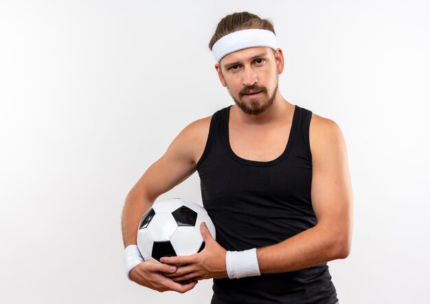 Уверенный молодой красивый спортивный мужчина в головной повязке и браслетах, держащий футбольный мяч, смотрит изолированно на белой стене