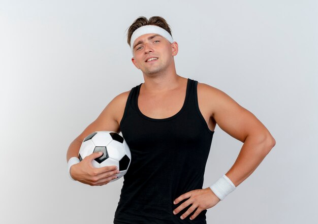 Confident young handsome sporty man wearing headband and wristbands holding soccer ball and putting hand on waist isolated on white 