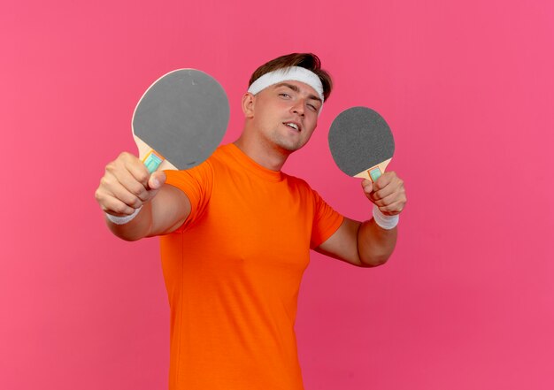 Confident young handsome sporty man wearing headband and wristbands holding and stretching out ping pong rackets isolated on pink  with copy space