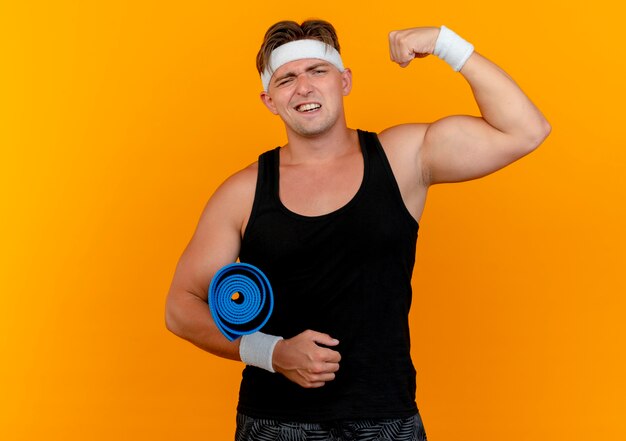 Confident young handsome sporty man wearing headband and wristbands holding towel and gesturing strong isolated on orange  with copy space