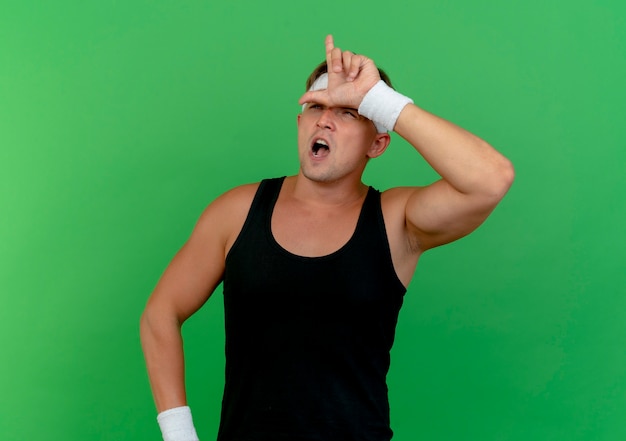 Free photo confident young handsome sporty man wearing headband and wristbands looking up and gesturing loser isolated on green