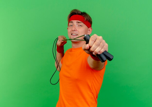 Confident young handsome sporty man wearing headband and wristbands pulling and stretching out jump rope towards camera isolated on green  with copy space