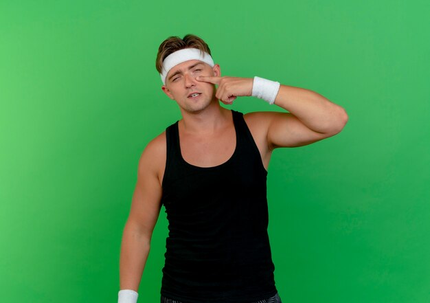 Confident young handsome sporty man wearing headband and wristbands putting finger under eye isolated on green  with copy space