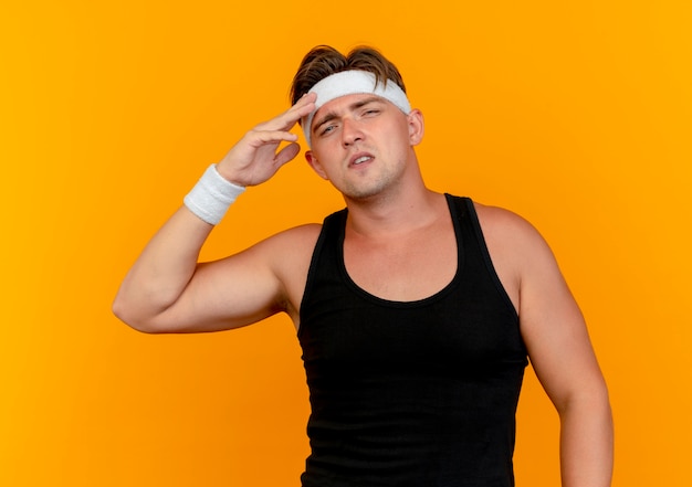 Confident young handsome sporty man wearing headband and wristbands putting hand on forehead isolated on orange 