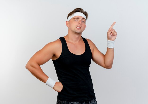 Confident young handsome sporty man wearing headband and wristbands putting hand on waist and pointing up isolated on white 