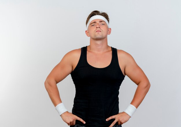 Confident young handsome sporty man wearing headband and wristbands putting hands on waist isolated on white 