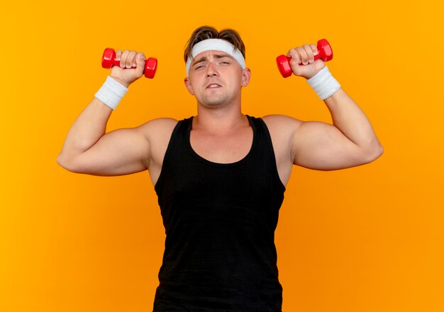 Confident young handsome sporty man wearing headband and wristbands raising dumbbells isolated on orange 