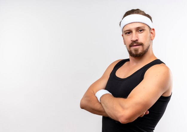 Confident young handsome sporty man wearing headband and wristbands standing with closed posture looking  isolated on white wall with copy space