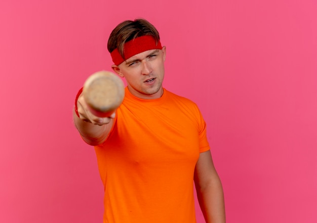 Confident young handsome sporty man wearing headband and wristbands stretching out baseball bat towards camera isolated on pink  with copy space
