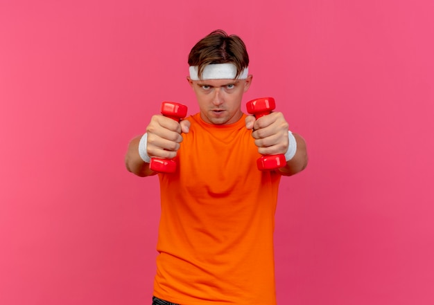 Free photo confident young handsome sporty man wearing headband and wristbands stretching out dumbbells isolated on pink  with copy space