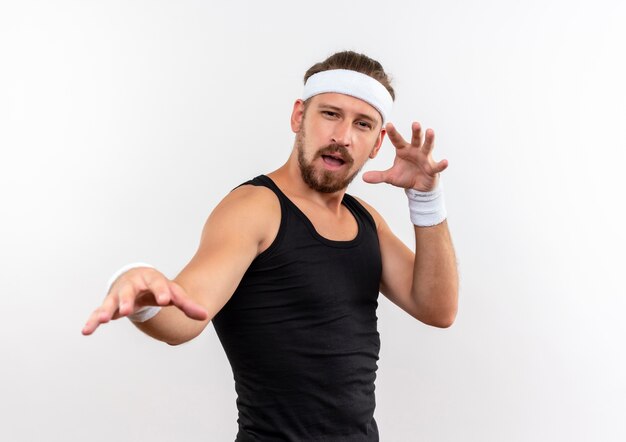 Confident young handsome sporty man wearing headband and wristbands stretching out hand and keeping another one near face isolated on white wall