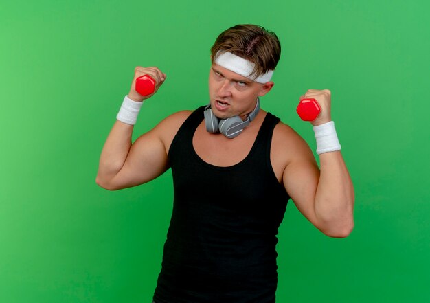 Confident young handsome sporty man wearing headband and wristbands with headphones on neck holding dumbbells looking straight isolated on green 