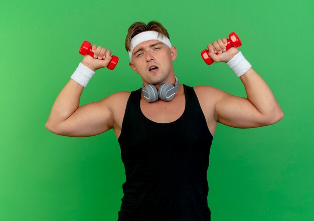 Confident young handsome sporty man wearing headband and wristbands with headphones on neck raising dumbbells isolated on green 