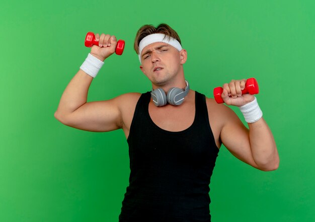 Confident young handsome sporty man wearing headband and wristbands with headphones on neck raising dumbbells isolated on green 