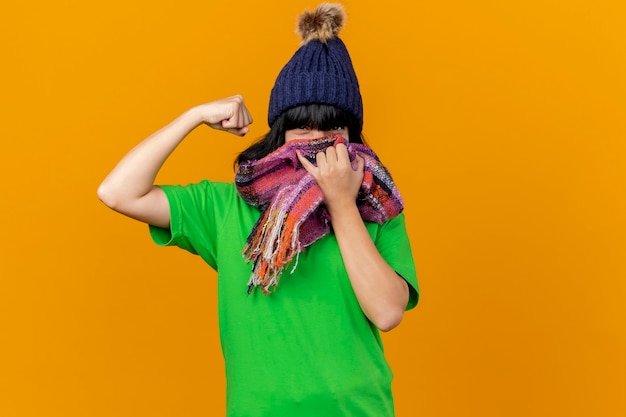 Confident young ill caucasian girl wearing winter hat and scarf covering mouth with scarf  doing strong gesture isolated on orange wall with copy space