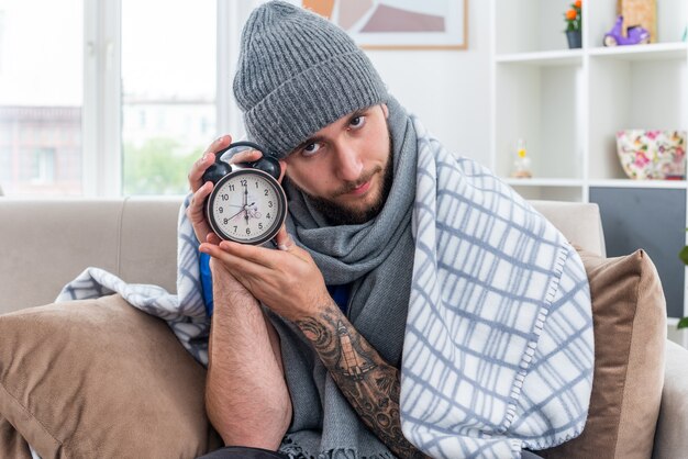 Confident young ill man wearing scarf and winter hat sitting on sofa in living room wrapped in blanket holding alarm clock looking at camera