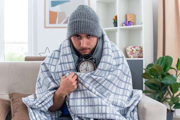 уверенный в себе молодой больной мужчина в шарфе и зимней шапке сидит на диване в гостиной, завернутый в одеяло, держит будильник и смотрит вперед