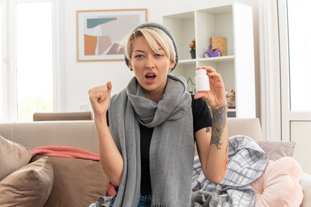 Confident young ill slavic woman with scarf around her neck wearing winter hat holding medicine bottle and keeping fist up sitting on couch at living room