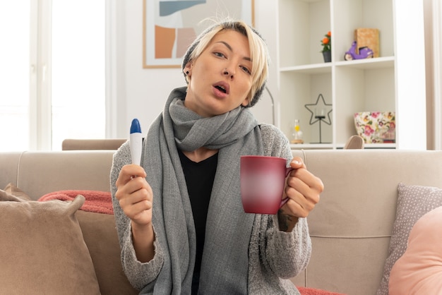 confident young ill slavic woman with scarf around her neck wearing winter hat holding thermometer and cup sitting on couch at living room