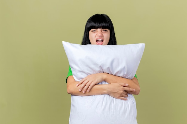 Уверенная молодая больная женщина обнимает подушку, глядя вперед, изолированную на оливково-зеленой стене